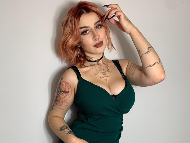 HannahSexy69 with red hair, green dress, tattoos, nose ring, choker, dark lipstick, holding hair, showing her tits, arm tattoos, and a confident smile.