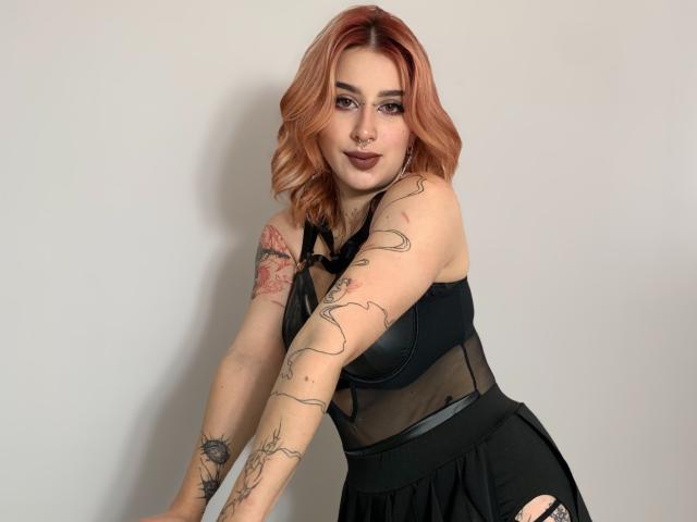 HannahSexy69 with pink hair, black mesh top, and tattoos, leaning forward, showing her tits and tattoos, with a septum ring and dark lipstick.