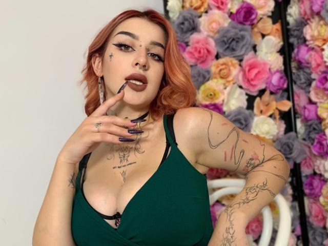 HannahSexy69 with pink hair, dark green bra, tattoos, and long nails, biting finger, green eyes, and a seductive look, showing her tits and a nose ring.