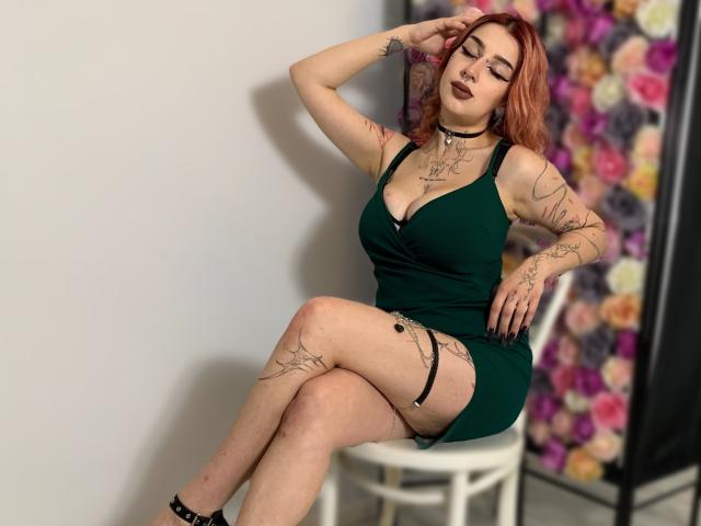 HannahSexy69 with red hair, green dress, tattoos, dark lipstick, sitting on white chair, legs crossed, hand in hair, black choker, garter, and black heels, eyes closed.