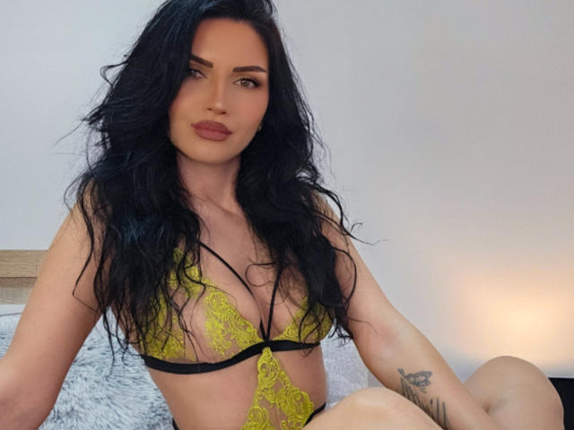 Paola Adams, con lunghi capelli neri, indossa un reggiseno di pizzo giallo e bretelle nere, mostrando il seno e un tatuaggio sul braccio destro, seduta su un letto..