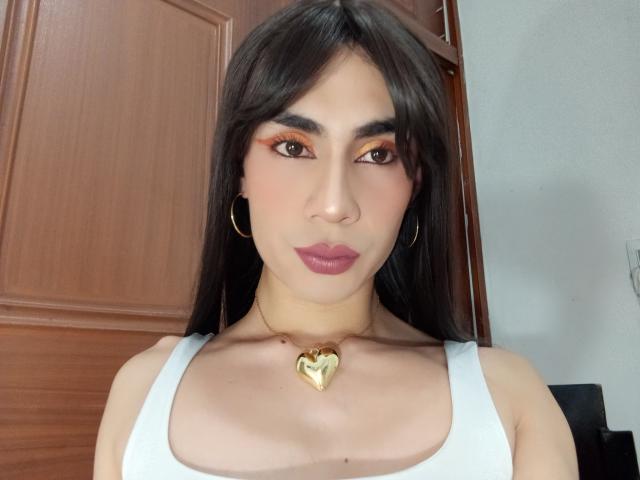 NiaaHills has long dark hair, orange eyeliner, pink lipstick, white tank top, gold heart necklace, gold hoop earrings, and a cute face with subtle makeup.