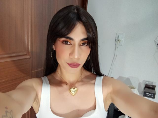 NiaaHills with dark hair, wearing a white tank top, gold heart necklace, and hoop earrings, showing her tits, armpits, and tattooed arm in a selfie.