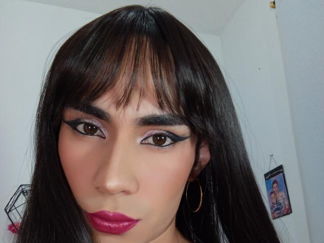 NiaaHills has dark brown hair, bangs, brown eyes with black eyeliner, pink eyeshadow, pink lipstick, and gold hoop earrings. Her skin is light brown, and she's looking at the camera.