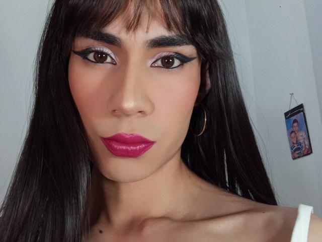 NiaaHills has a cute face with dark straight hair, pink lipstick, black winged eyeliner, and a small mole on her neck. She's wearing a white top and gold hoop earrings.