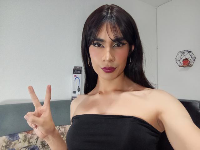 NiaaHills with dark hair, makeup, and purple lipstick, showing a peace sign with her right hand, wearing a black strapless top, her small tits visible.