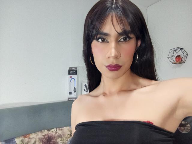 NiaaHills with long black hair, hoop earrings, and dark purple lipstick, showing bare shoulders and black strap, exposing a small red nipple. NiaaHills has a sexy, confident look.