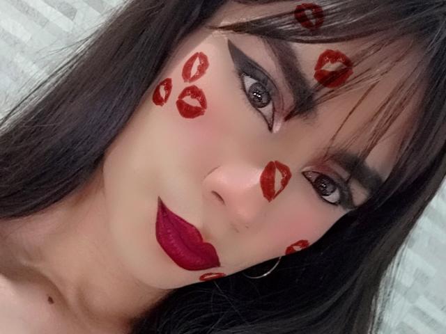 NiaaHills has dark makeup, red lipstick, and red kiss marks on her face. She has black hair, hoop earrings, and her eyes are looking at the camera.