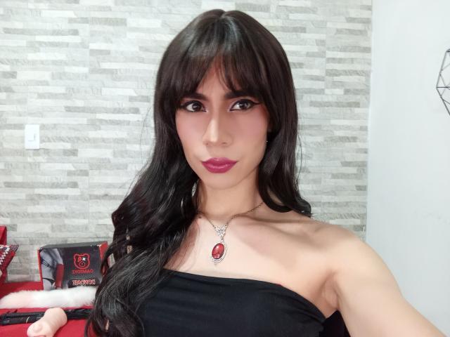 NiaaHills with long black hair, dark pink lipstick, strapless black top, necklace with red gem, holding red and black bag, light skin, brown eyes, standing in front of textured wall.