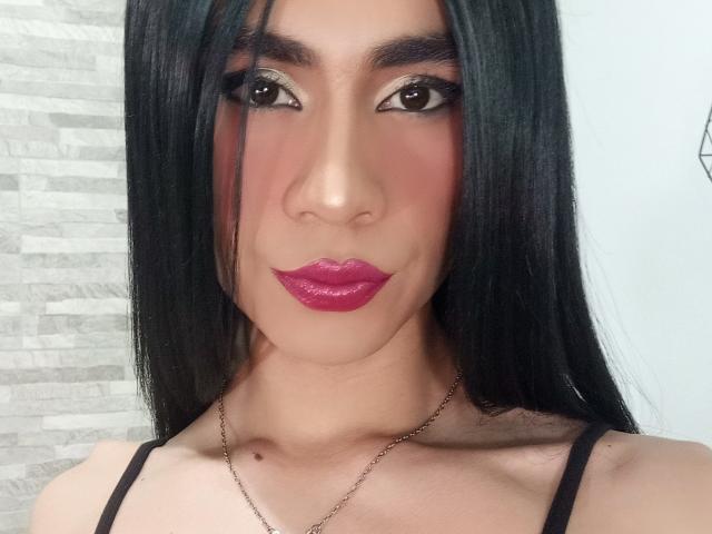 NiaaHills has long black hair, shiny pink lipstick, dark makeup, and a cute smile. She's wearing a black top with a thin chain necklace, showing her tits.