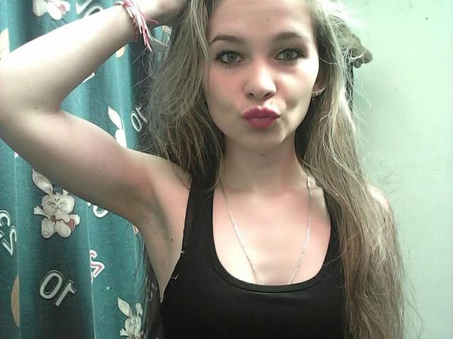 DomenikHot with long grayish-brown hair, pouty pink lips, black tank top, silver necklace, and arm raised, wearing pink bracelets, looking at camera.