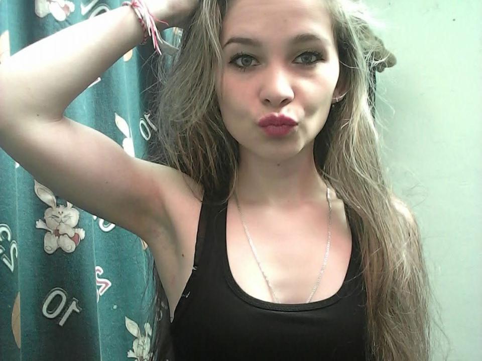 DomenikHot with long grayish-brown hair, pouty pink lips, black tank top, silver necklace, and arm raised, wearing pink bracelets, looking at camera.