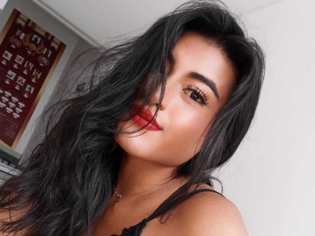 MichelleDenise with long black hair, red lipstick, and thick eyelashes, her wavy hair partially covering her eye, wearing a black strap top, her ass slightly visible.