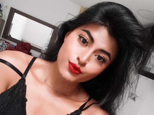 MichelleDenise has shiny black hair, red lipstick, and is wearing a black lace top with thin straps, looking at the camera with brown eyes. She's also wearing a delicate necklace.