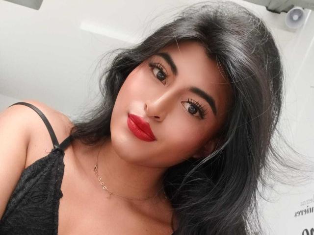 MichelleDenise has long black hair, red lipstick, and thick eyebrows, wearing a black lace top, with a gold necklace, looking at the camera with a slight smile.