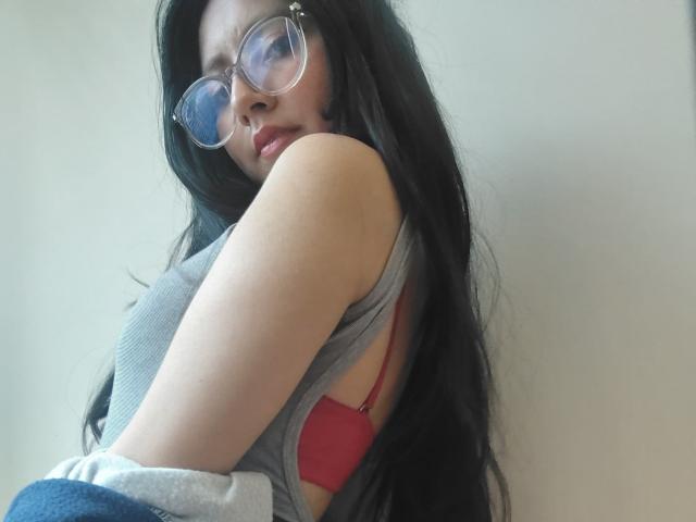 VioletXBloom with long black hair, glasses, gray tank top, red bra, looking over shoulder, showing her tits, light skin, soft expression, casual pose, slightly blurred background.
