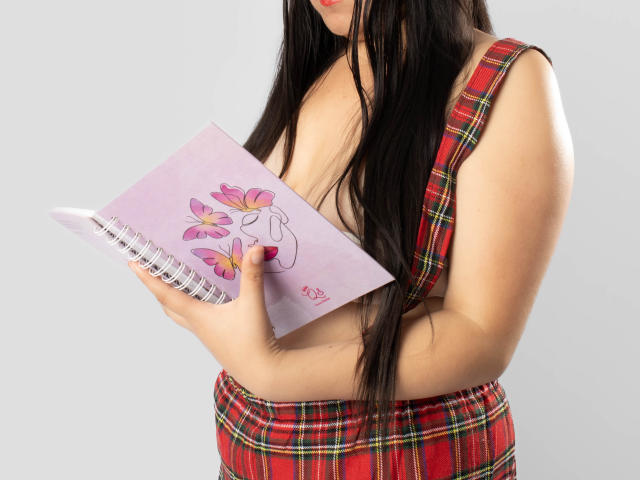 SaraFountain with long black hair, wearing a red plaid dress, holding a pink notebook with pink flower doodles, showing her big boobs, in a photograph.