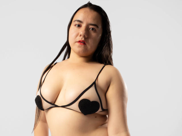 SaraFountain with long black hair, wearing a sheer black bra with heart pasties covering her tits, standing against a white background, looking directly at the camera.