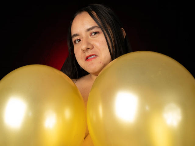 SaraFountain with shiny yellow balloons in front, black hair, red lipstick, and a slight smile, holding them up to her chest.