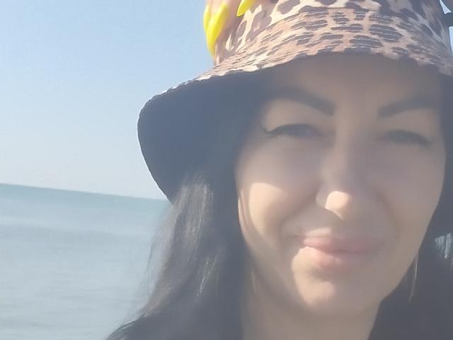 MirandaHott69 with a leopard-print bucket hat, smiling, has long black hair, and is standing by a blue ocean, wearing yellow ear accessories, with a light skin tone.