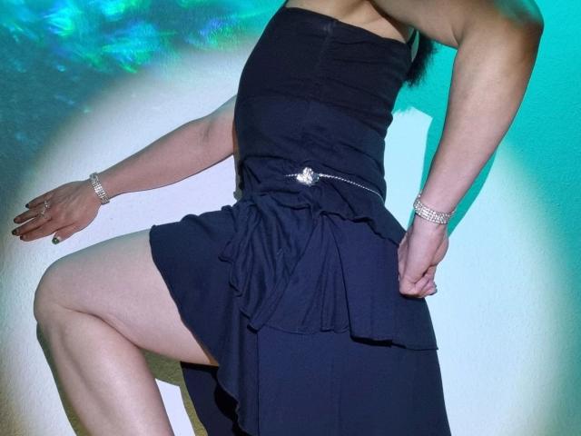 MelissaSin-hot wearing a strapless black dress, silver bracelet, and ring, knee raised, showing thigh, with a silver chain belt, arm raised, blue-green light, visible arm muscles.