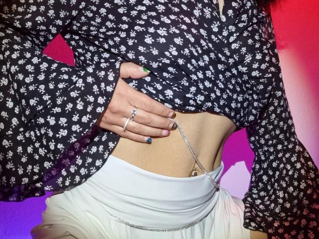 MelissaSin-hot's hand on her bare, flat midriff, wearing a black floral shirt, white skirt, and multiple rings, with a belly button piercing and green nails.