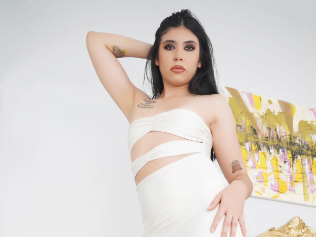 MiluSky with black hair, wearing a white strapless dress, has tattoos on her arm and chest, standing with one arm behind her head, exposing her tits.