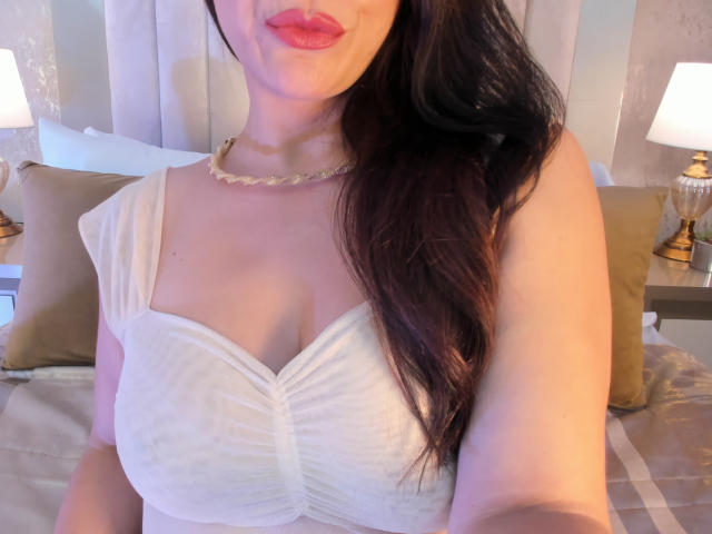 CameronViviane, com cabelos longos e escuros, batom rosa, blusa branca sem alças que mostra seios grandes, decote discreto, colar, sentada na cama com travesseiros e luminárias ao fundo..