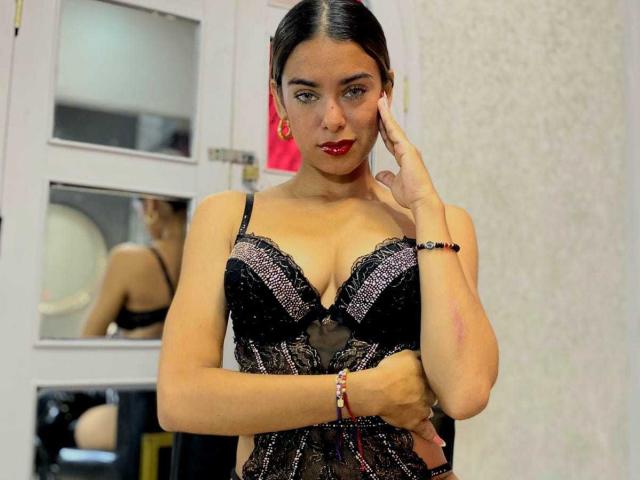 VanelopeRusso with dark hair, red lipstick, wearing black lace lingerie, showing her tits, hand on cheek, looking at camera, reflection in mirror, white wall background.