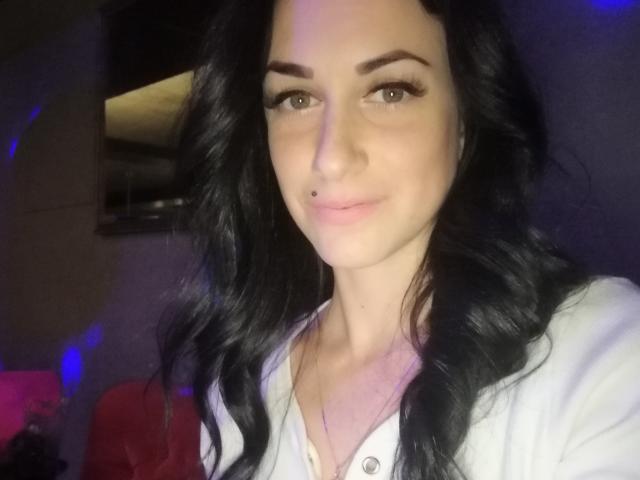 MiaDefne has black wavy hair, light skin, green eyes, and a small nose piercing. She's wearing a white shirt, looking at the camera with a subtle smile.