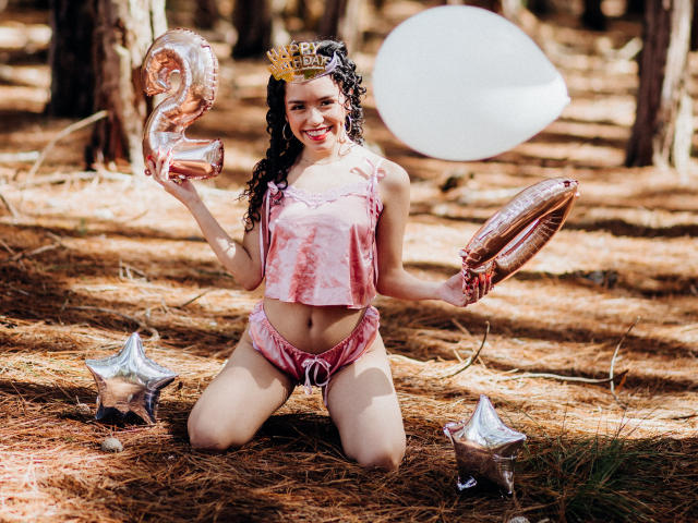 Macaroux kneeling on pine needles, wearing pink tie-dye crop top and shorts, holding metallic balloons, crown, smiling, curly black hair, forest background, white balloon in air.