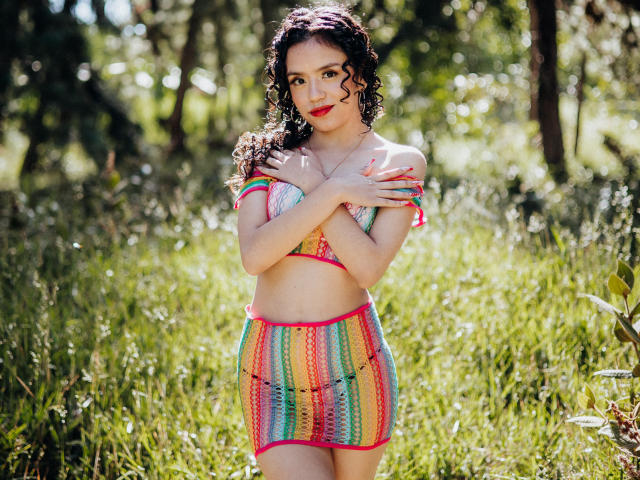 Macaroux with curly dark hair, wearing a colorful crochet bikini top and skirt, standing in grass, arms crossed over small tits, red lipstick, and a cute smile.