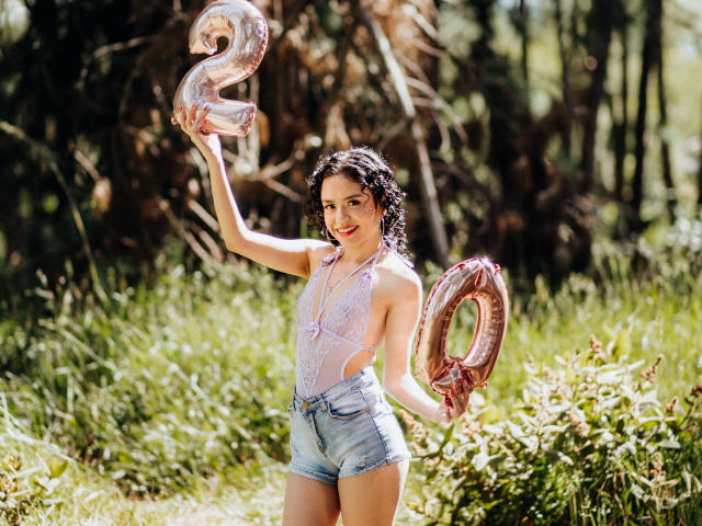 Macaroux with curly black hair, wearing a pink lace bodysuit and denim shorts, holding shiny silver "20" balloons, smiling in a sunlit grassy area.