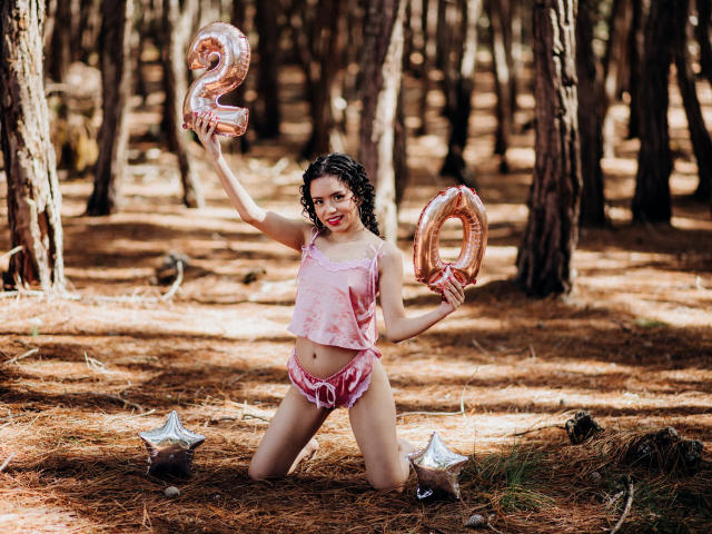 Macaroux with curly black hair, pink tie-dye crop top, and pink shorts, kneeling in forest, holding pink metallic "2" and "0" balloons, on pine-covered ground.