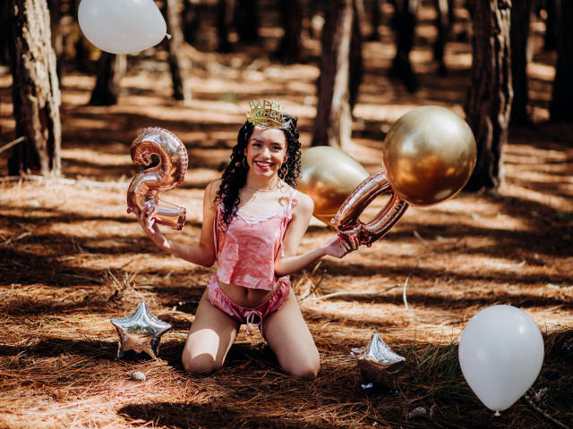 Macaroux kneels in a forest, wearing a pink lace top and shorts, gold crown, holding metallic balloons, white balloons, and star-shaped decorations, smiling with curly dark hair.