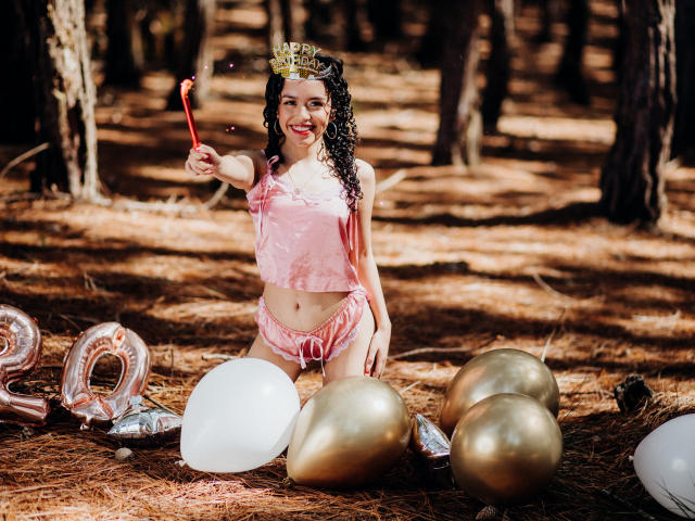 Macaroux com cabelo cacheado, blusa rosa de veludo e shorts, coroa dourada, segurando uma varinha vermelha, sorrindo, ajoelhada sobre agulhas de pinheiro, cercada por balões dourados e brancos..