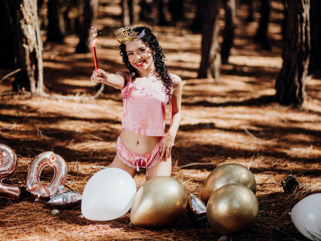 Macaroux with curly black hair, pink velvet top, and matching shorts, wearing a gold crown and holding a sparkler, kneeling in a forest with balloons and metallic decor.