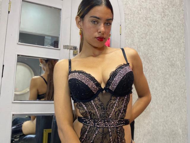 Vanelope Russo, con i capelli castano scuro raccolti in una coda di cavallo, indossa un abito intimo in pizzo nero e rosa, che mette in mostra il seno e il sedere. È di fronte a uno specchio..