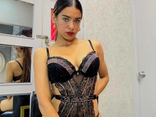 VanelopeRusso with dark hair, red lipstick, wearing black lace lingerie with sparkling details, showcasing her tits and slim waist, standing in a room with mirrors.