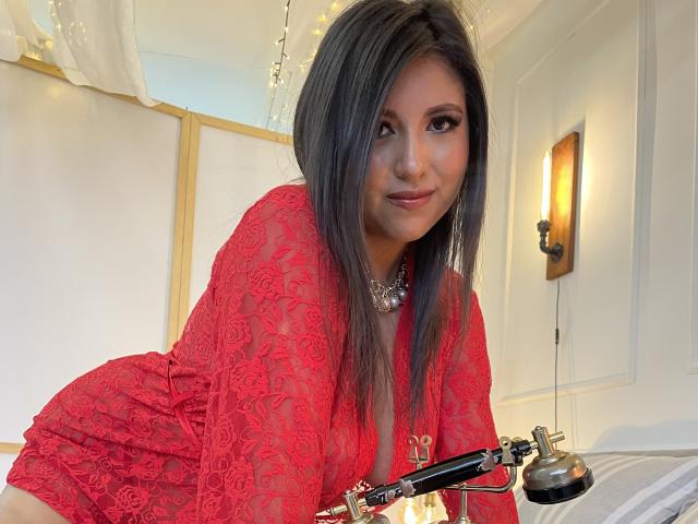 EvangelineAx with long dark hair, red lace dress, and necklace, leaning forward, smiling, showing her tits, holding a silver lamp, in a brightly lit room.