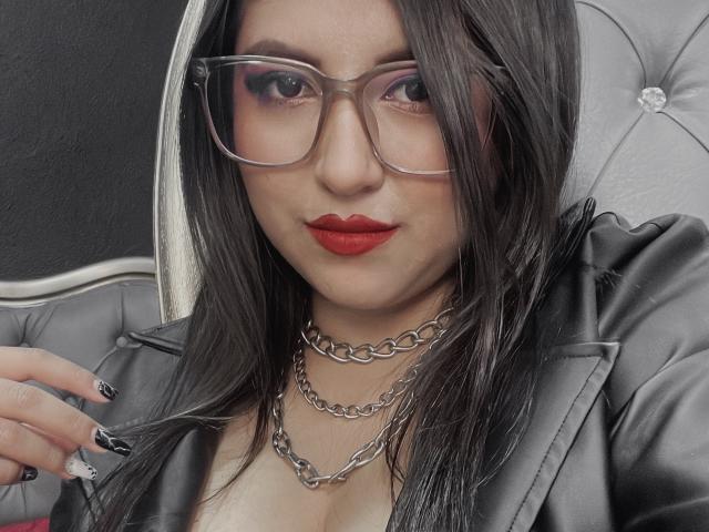 EvangelineAx with glasses, red lipstick, black nails, silver chain necklaces, and a grey leather jacket, looking at the camera with a slight smile. Boobs visible.