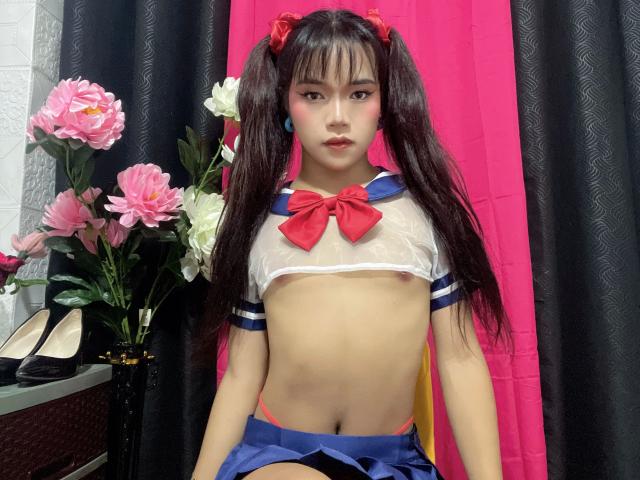 FRESHotBHEAx-hot with long black pigtails, wearing a sheer white top with red bow, exposing small tits, and a blue skirt, standing in front of pink and black curtains.