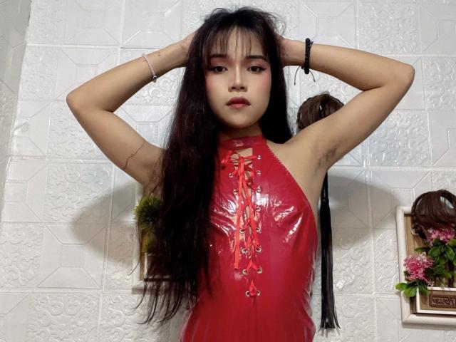 FRESHotBHEAx-hot with long black hair, wearing a shiny red PVC dress, hands in her hair, armpit hair visible, looking at camera, red lipstick, white tiled wall.
