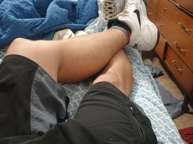 DirtyLegforYou's legs crossed at the ankle, wearing black shorts and white Nike sneakers with socks, lying on a blue patterned bed, next to a wooden dresser.