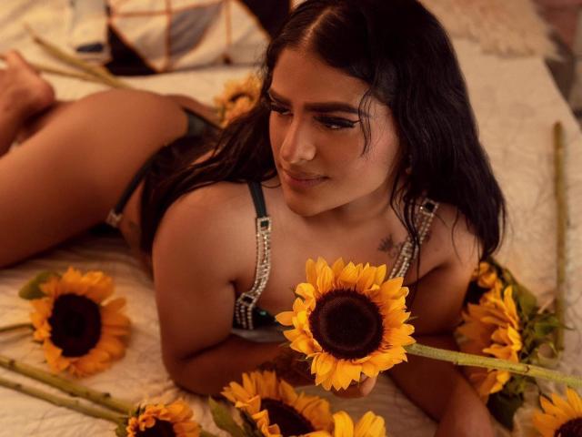PartyNasty, con piel morena, cabello negro y tatuaje, acostada en la cama, vistiendo un sujetador verde y blanco, sosteniendo un girasol, rodeada de girasoles, mostrando el trasero, mirando hacia arriba con una sonrisa sutil..
