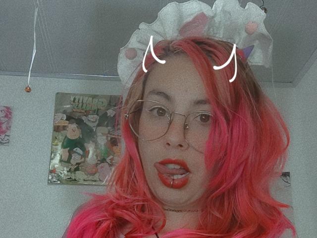 NatashaLau mit rosa Haaren, rotem Lippenstift und Katzenohren, die eine Brille trägt, hat ein attraktives Gesicht, wobei ihre Zunge leicht herausragt. Ihr Hintern ist nicht sichtbar..