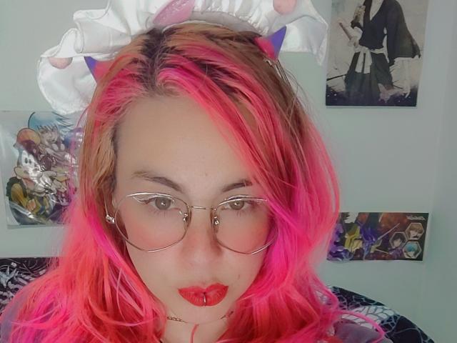 NatashaLau with pink and blonde hair, wearing glasses, red lipstick, and a white headband, looking at the camera with a slight pout. Boobless, no visible ass or pussy.