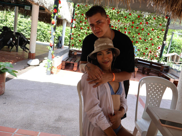 AriandDante with small tits and blue bra, standing in front of AriandDante with short brown hair, black shirt, and tan hat, under a thatched roof, holding her arm.