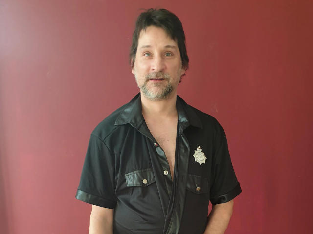 Mario41-hot, middle-aged dude with a slightly grubby beard, wearing a black leather shirt with a police badge, standing against a red wall, looking relaxed and slightly goofy.