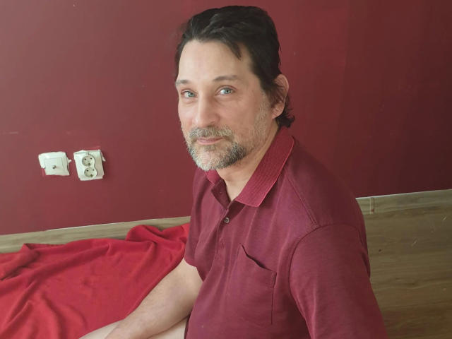 Mario41-hot has a slightly graying beard, short brown hair, and is wearing a red polo shirt. He's sitting on a red cloth with a wooden floor.